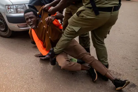 BADRU KATUMBA / AFP Ugandan police arrest an environmental activist during a protest in Kampala against the East African Crude Oil Pipeline Project. The protestor is on the floor and has his mouth open, as if he is shouting, while members of the police grip his shirt.