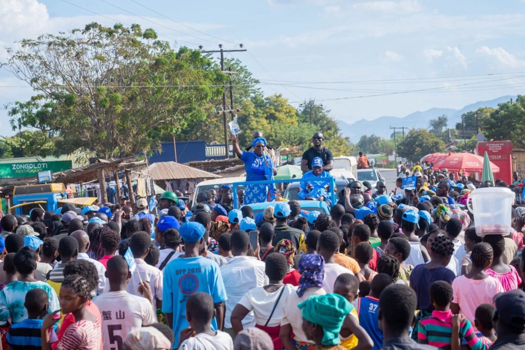 A large crowd gathers in a busy street to attend a political rally, with people wearing blue Democratic Progressive Party (DPP) clothing and caps. Two party representatives, dressed in blue attire, stand on a vehicle addressing the audience, while others capture the moment on phones. The event takes place in a small-town setting with shops, trees, and mountains visible in the background.