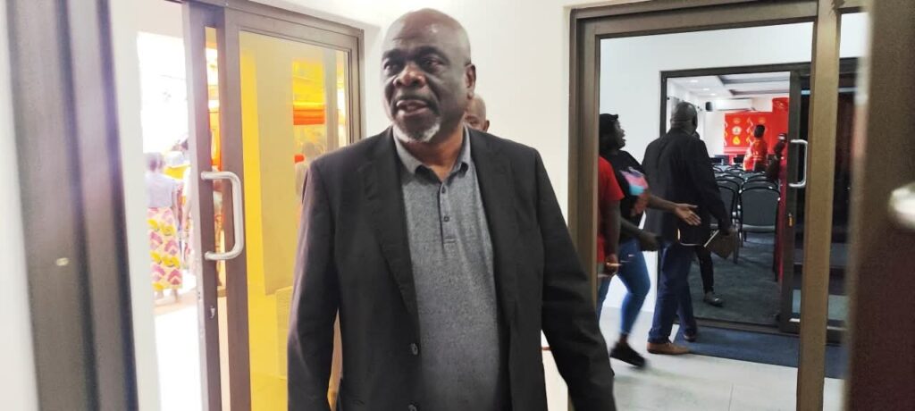 A middle-aged man wearing a dark blazer over a grey polo shirt walks through a doorway in an indoor setting. Behind him, several people are visible walking or standing in a hallway and adjacent room, some wearing casual clothing, with chairs and a red banner visible in the background.

