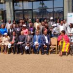 a large group of people, including men and women, some in wheelchairs, pose for a group photo outside a building with glass doors. several people are seated in front, and others stand behind them. a banner is visible on the right.