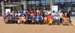 a large group of people, including men and women, some in wheelchairs, pose for a group photo outside a building with glass doors. several people are seated in front, and others stand behind them. a banner is visible on the right.