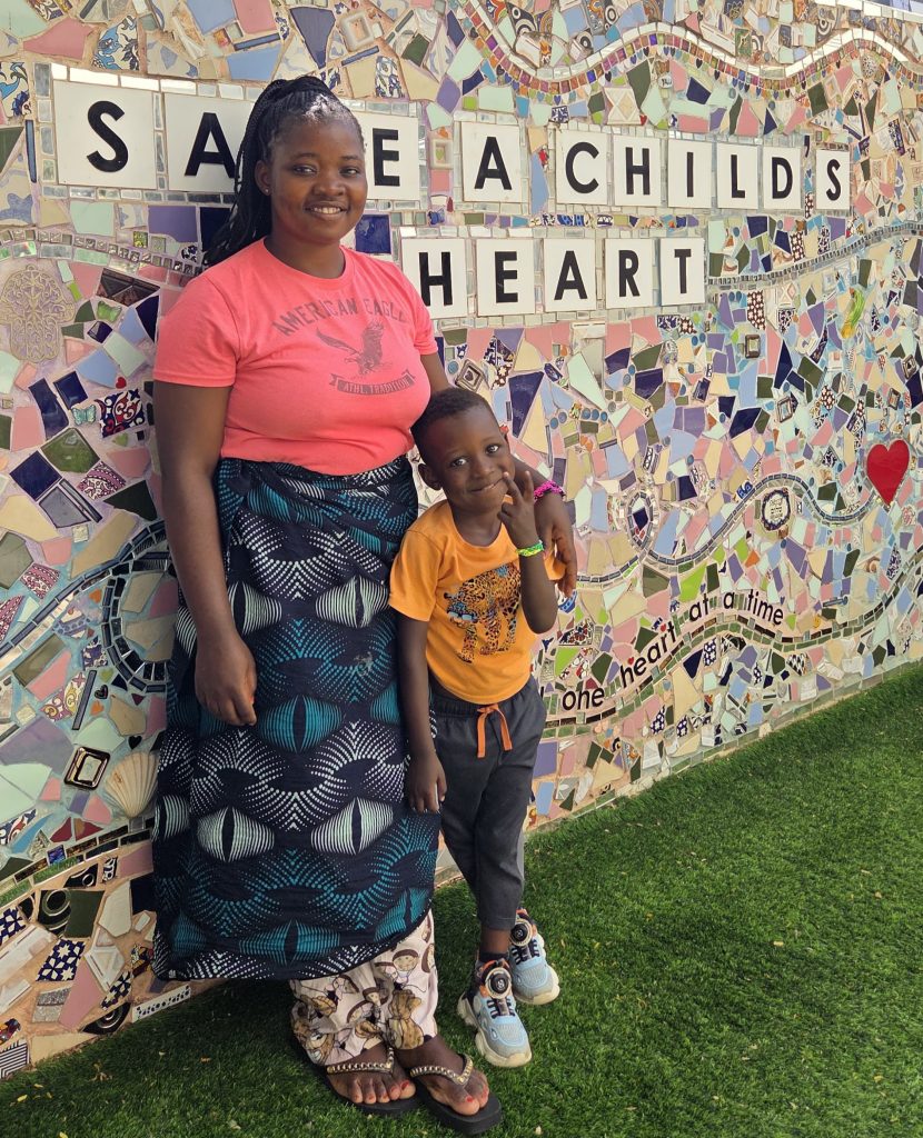 Magret Pitani and her son Prince stand in front of a mosaic wall reading Save a Child's Heart