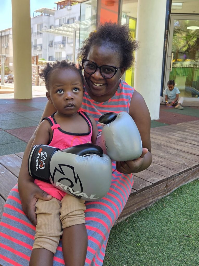A smiling Malawian mother holds her young daughter, who is wearing oversized boxing gloves