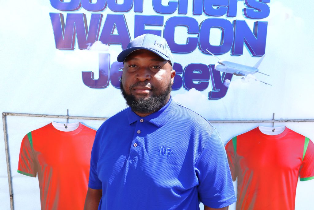 NBM plc Products Development and Strategy Manager Ibrahim Chapeyama stands in front of a Waku WAFCON promotion backdrop with Scorchers jerseys displayed behind him